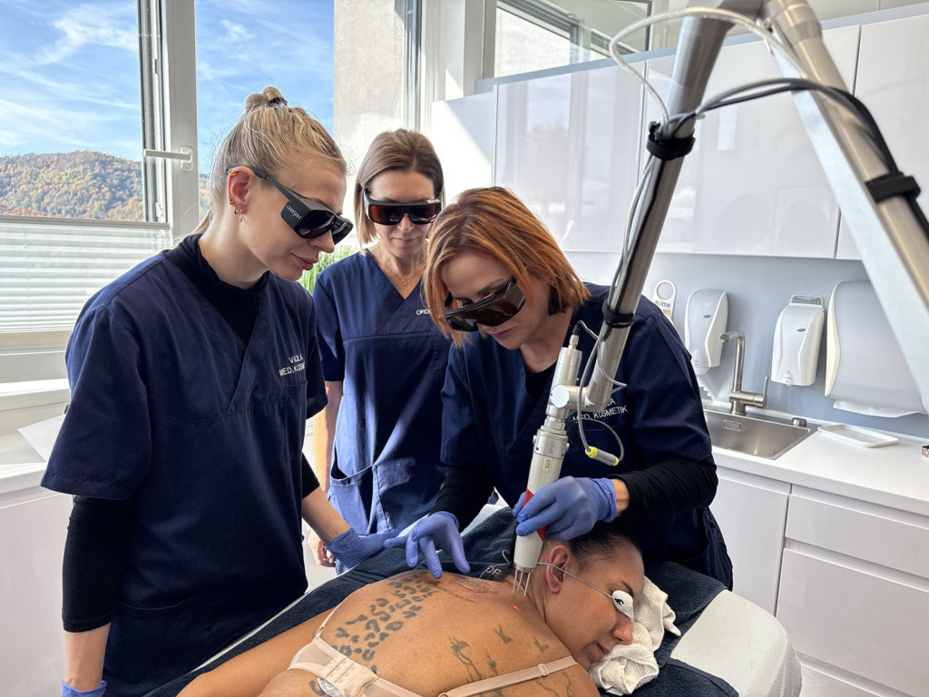 Dame liegt auf dem Bauch auf einer Liege und lässt mit einem Laser ein Tattoo-Entfernung. Neben ihr stehen drei Damen, die die Tattoo-Entfernung mit dem Laser durchführen